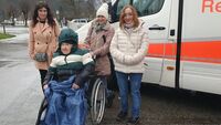 Drei Frauen stehen mit einem älteren Mann im Rollstuhl vor einem Krankenwagen.