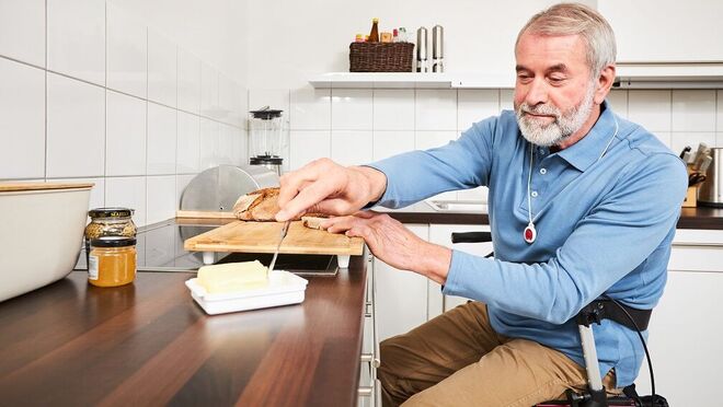 Ein Mann mit einem Hausnotrufgerät am Handgelenk sitzt auf einem Rollator an der Küchenzeile und streicht sich ein Brot.